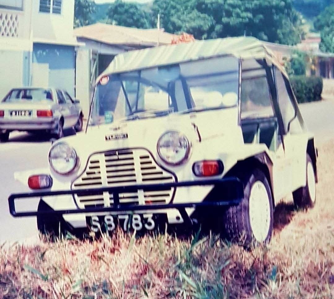 Mahé'de bir sokakta eski bir sedanın ve dükkânın önünde park etmiş krem rengi branda tavanlı Mini Moke